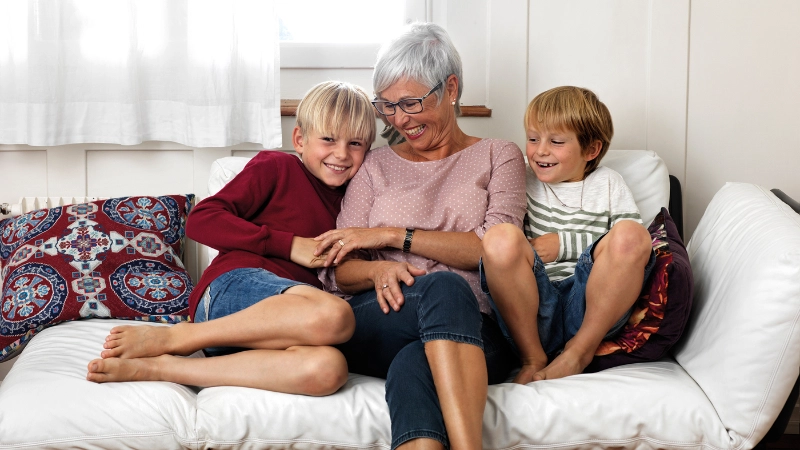 Une femme âgée est assise sur un canapé avec ses deux petits-enfants et ils rient.